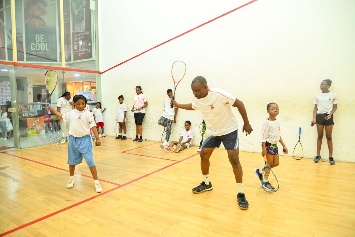 Coach demonstrating squash technique to a young participant at the Ikoyi Club Squashtival Summer Camp, with other children watching from the court walls