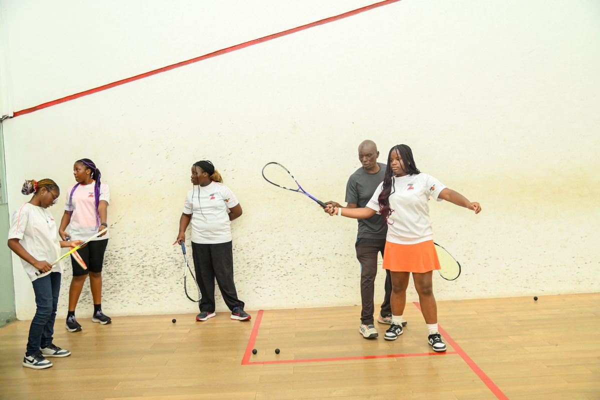 Coach physically guiding a girl's swing technique while three other participants watch during the Ikoyi Club Squashtival Summer Camp
