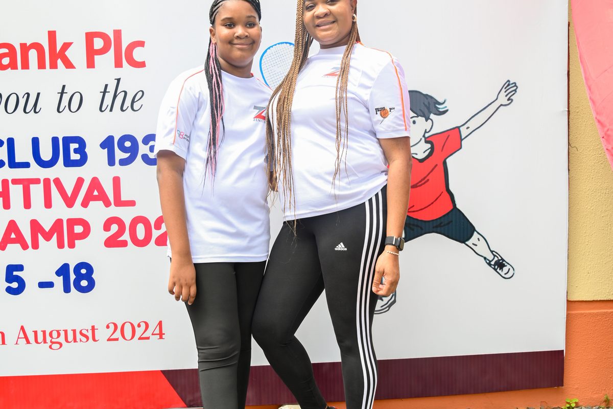 Two young female participants posing in front of the official Ikoyi Club 1938 Squashtival Summer Camp programme backdrop